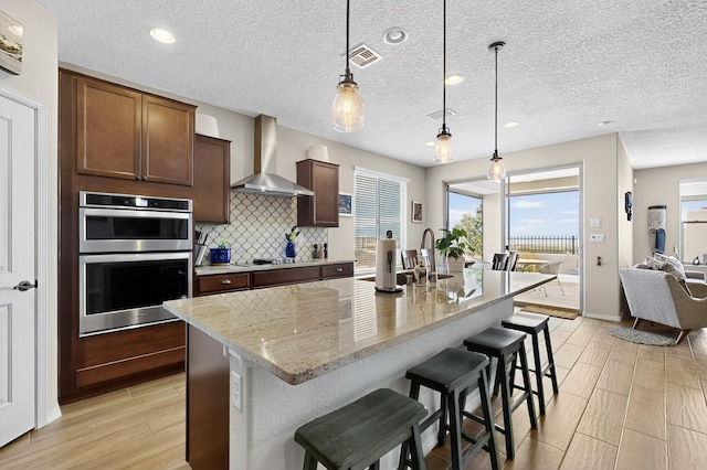 6417 Kings Cyn NE, Rio Rancho, NM 87144 - kitchen with wall chimney range hood, stainless steel double oven, tasteful backsplash, light wood-style floors, and a breakfast bar area