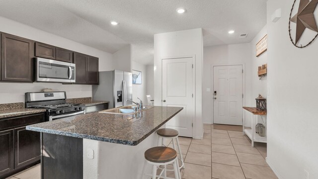 2404 Del Timbre Ln SW, Albuquerque, NM 87121 - kitchen featuring appliances with stainless steel finishes, an island with sink, a kitchen breakfast bar, dark brown cabinetry, and light tile patterned floors