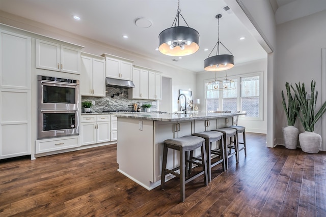 kitchen featuring decorative light fixtures, white cabinetry, double oven, and a center island with sink