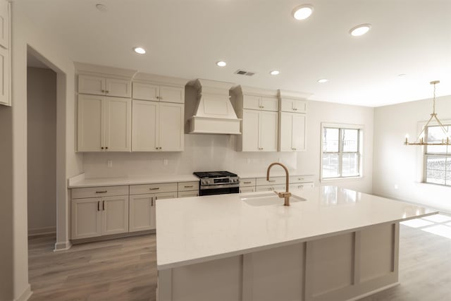 kitchen with pendant lighting, sink, premium range hood, an island with sink, and stainless steel range with gas cooktop