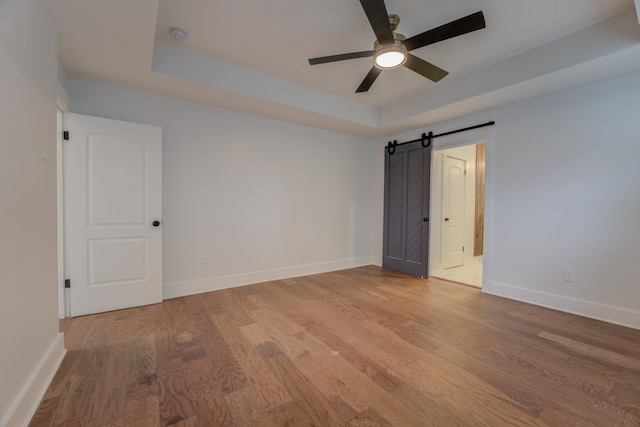 unfurnished room featuring a barn door, wood-type flooring, ceiling fan, and a tray ceiling