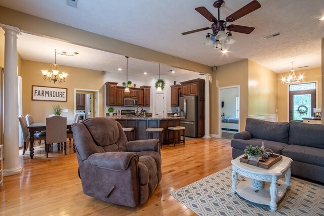 living room with light wood-type flooring, ceiling fan with notable chandelier, sink, ornate columns, and a textured ceiling