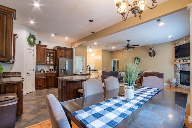 tiled dining room featuring ceiling fan with notable chandelier, sink, and a stone fireplace