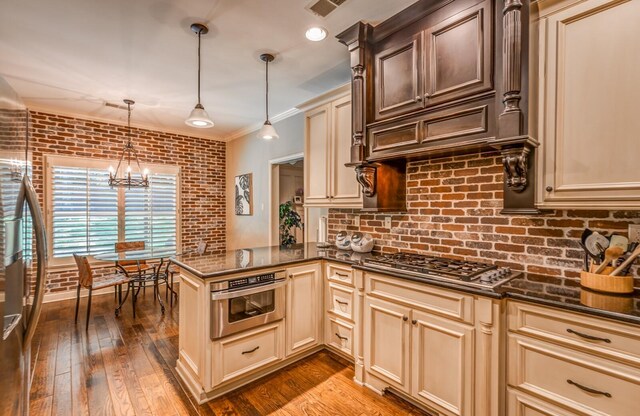 kitchen with cream cabinetry, a notable chandelier, stainless steel appliances, brick wall, and a peninsula