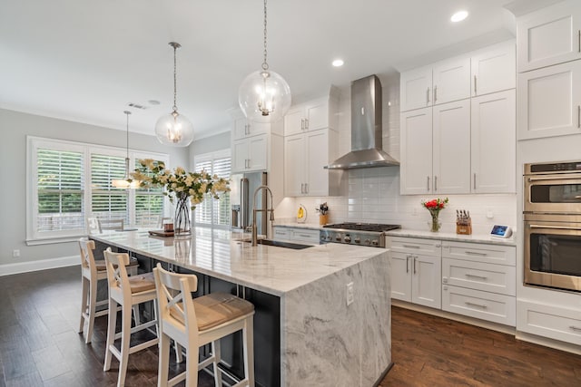 kitchen with a kitchen island with sink, white cabinets, hanging light fixtures, and wall chimney range hood