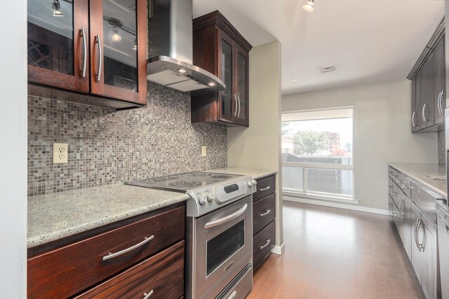 kitchen featuring dark brown cabinetry, light stone counters, stainless steel electric range oven, decorative backsplash, and wall chimney exhaust hood