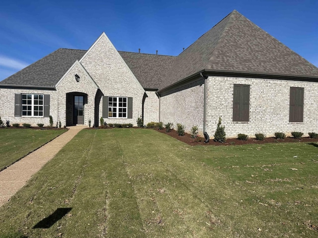 french country home with a shingled roof, a front yard, and brick siding