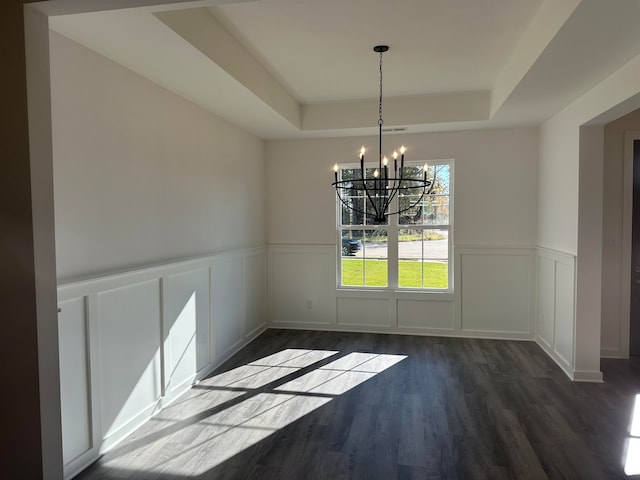 unfurnished dining area with a raised ceiling, dark wood finished floors, a wainscoted wall, a chandelier, and a decorative wall