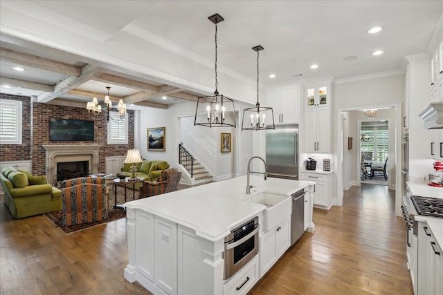 kitchen with white cabinets, decorative light fixtures, stainless steel appliances, an island with sink, and a brick fireplace