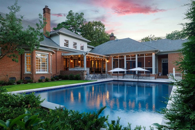 back of property at dusk with brick siding, a sunroom, an outdoor pool, a chimney, and a patio area