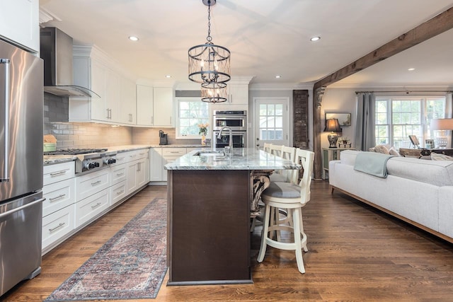 kitchen featuring white cabinetry, wall chimney exhaust hood, stainless steel appliances, light stone counters, and a kitchen island with sink