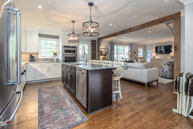 kitchen featuring stainless steel appliances, dark stone counters, pendant lighting, a kitchen island with sink, and white cabinets