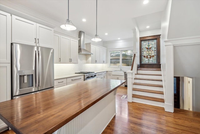 kitchen featuring wooden counters, wall chimney exhaust hood, stainless steel appliances, pendant lighting, and white cabinets