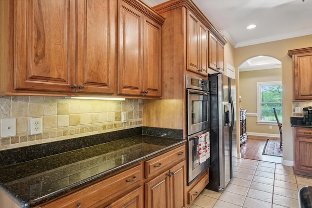 kitchen featuring light tile patterned floors, crown molding, appliances with stainless steel finishes, dark stone countertops, and backsplash