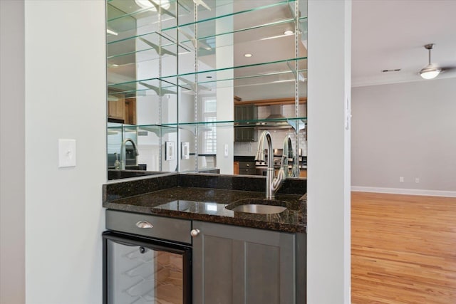 bar with sink, light hardwood / wood-style flooring, dark stone countertops, ornamental molding, and beverage cooler