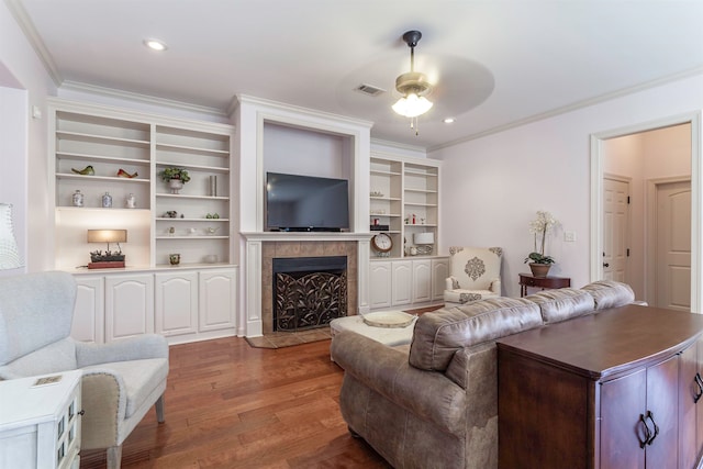 living room featuring ceiling fan, built in features, dark hardwood / wood-style floors, a tile fireplace, and crown molding