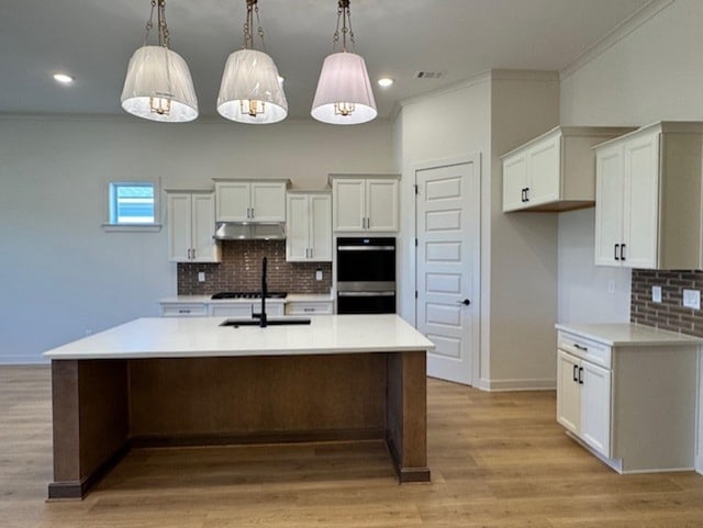 kitchen with light wood-type flooring, stainless steel double oven, a kitchen island with sink, pendant lighting, and white cabinetry