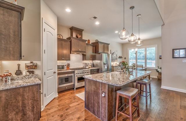 kitchen featuring appliances with stainless steel finishes, a kitchen island with sink, light stone countertops, decorative light fixtures, and wood-type flooring