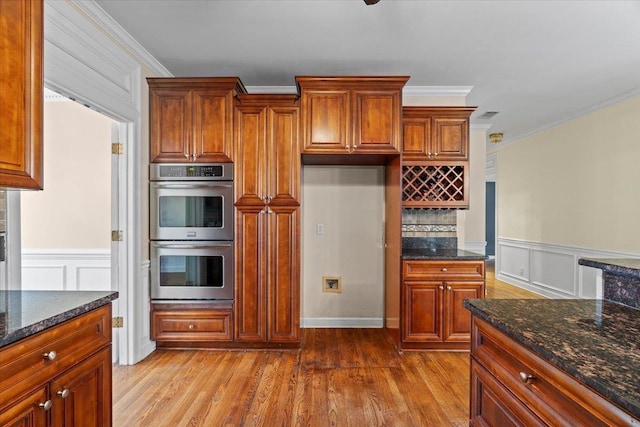 kitchen featuring brown cabinets, crown molding, stainless steel double oven, wood finished floors, and dark stone counters