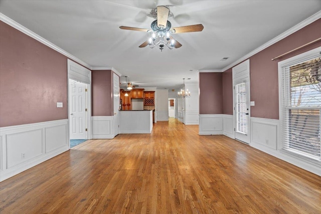 unfurnished living room with ornamental molding, a wainscoted wall, light wood finished floors, and ceiling fan with notable chandelier