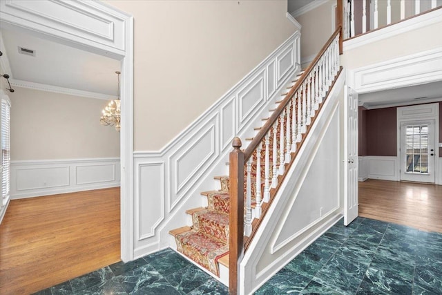 stairs with a notable chandelier, a wainscoted wall, wood finished floors, visible vents, and ornamental molding