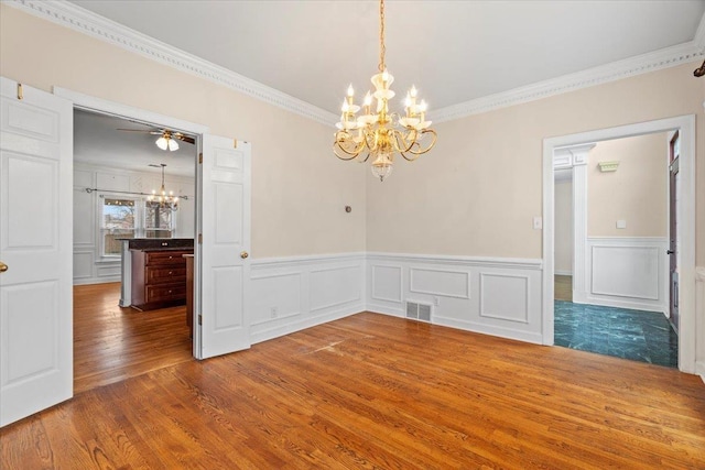 empty room with visible vents, wainscoting, ornamental molding, wood finished floors, and ceiling fan with notable chandelier