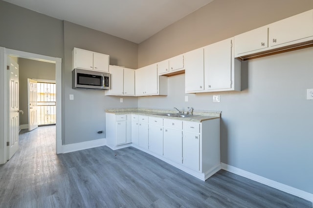 kitchen with a sink, stainless steel microwave, wood finished floors, white cabinets, and baseboards