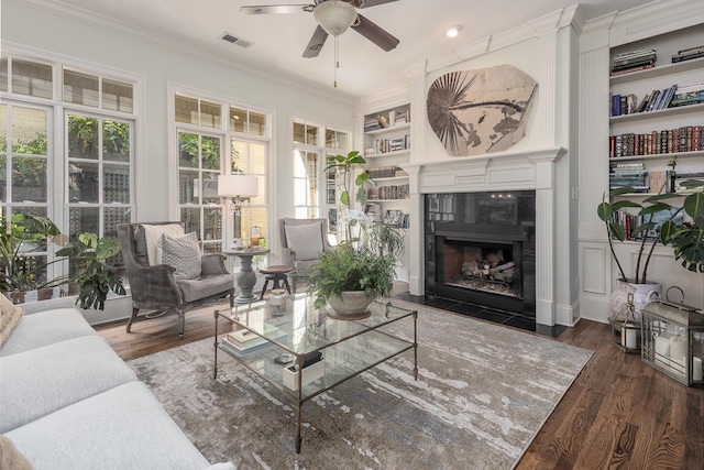 living room featuring a tiled fireplace, crown molding, ceiling fan, built in shelves, and dark hardwood / wood-style floors