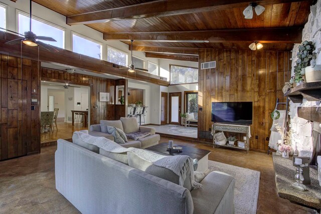 living room featuring beamed ceiling, high vaulted ceiling, wooden walls, a stone fireplace, and ceiling fan