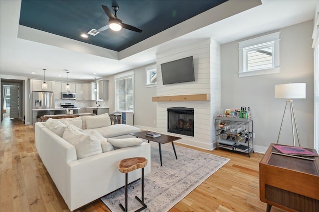 living room with visible vents, light wood finished floors, a fireplace, a ceiling fan, and a tray ceiling