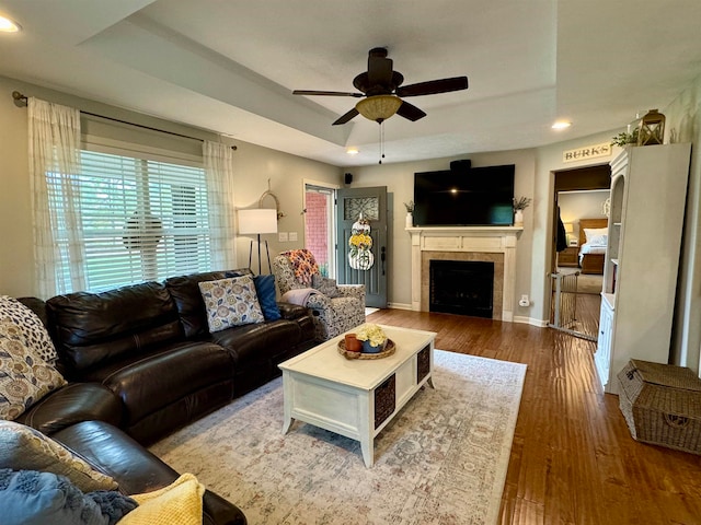 living room featuring wood-type flooring, a tray ceiling, a tiled fireplace, and ceiling fan