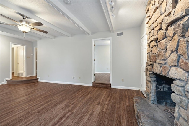 unfurnished living room featuring ceiling fan, beamed ceiling, a stone fireplace, a textured ceiling, and dark hardwood / wood-style flooring