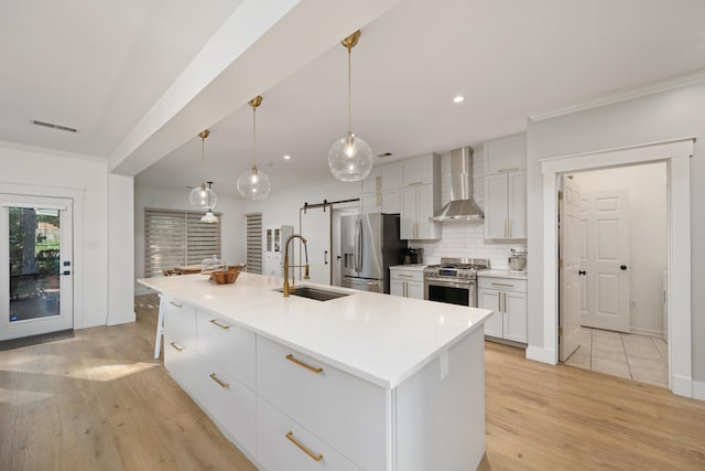 kitchen featuring light hardwood / wood-style floors, an island with sink, wall chimney range hood, a barn door, and appliances with stainless steel finishes