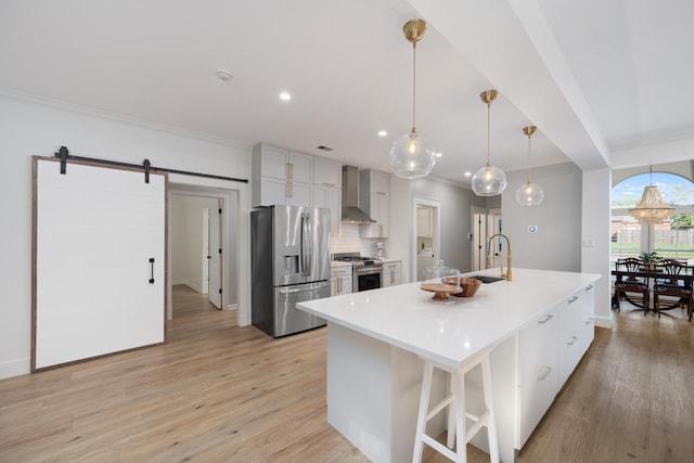 kitchen featuring a kitchen island with sink, a barn door, wall chimney exhaust hood, stainless steel appliances, and decorative light fixtures