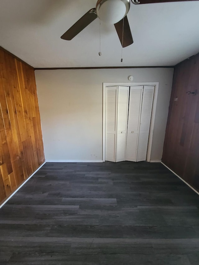 unfurnished bedroom featuring a closet, dark wood-type flooring, wooden walls, crown molding, and ceiling fan