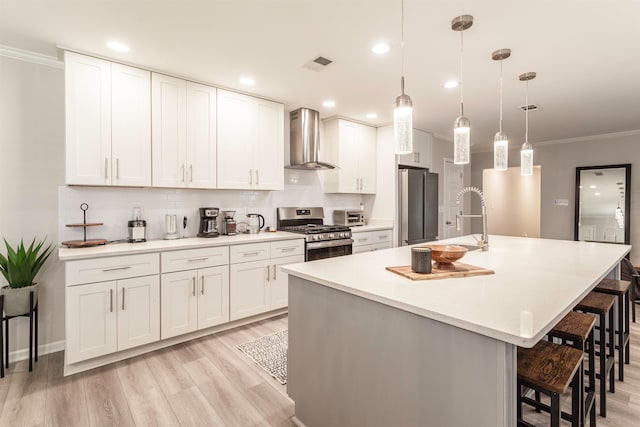 kitchen with white cabinets, an island with sink, wall chimney exhaust hood, decorative light fixtures, and appliances with stainless steel finishes