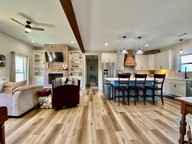 living room with ceiling fan, a brick fireplace, beam ceiling, and light hardwood / wood-style floors