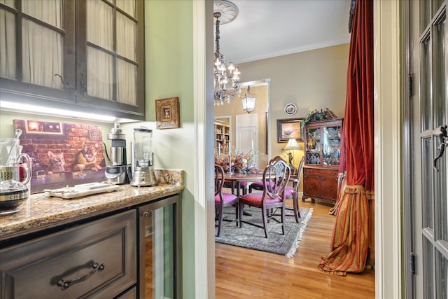 bar with light stone counters, ornamental molding, beverage cooler, a chandelier, and light wood-type flooring