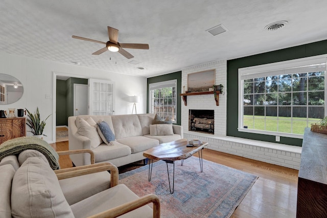 living room featuring a brick fireplace, light hardwood / wood-style flooring, a textured ceiling, and ceiling fan