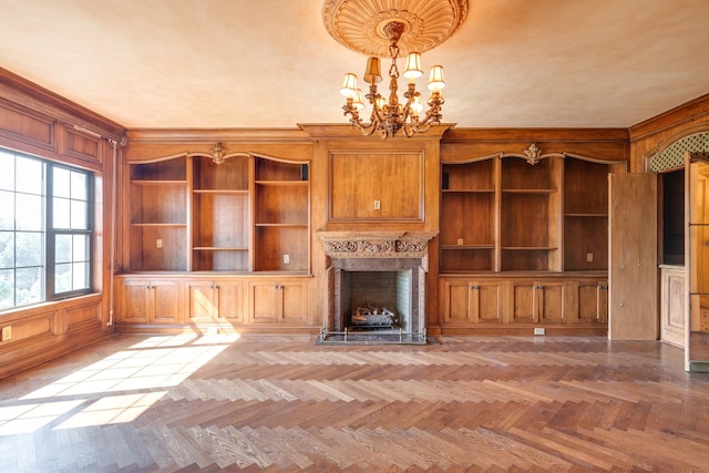 unfurnished living room with wooden walls, light parquet floors, a notable chandelier, a high end fireplace, and built in shelves