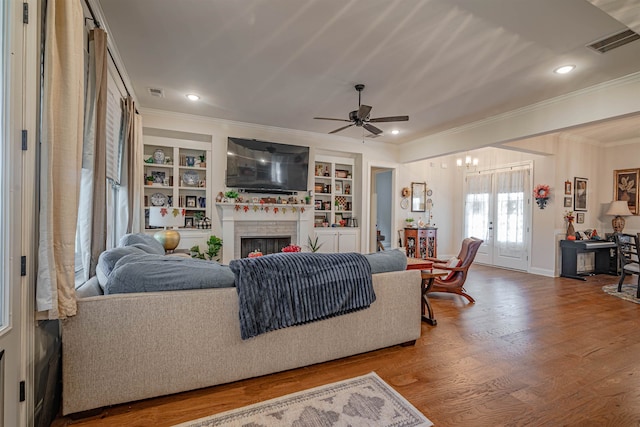 living room with ceiling fan, wood-type flooring, a brick fireplace, built in shelves, and crown molding