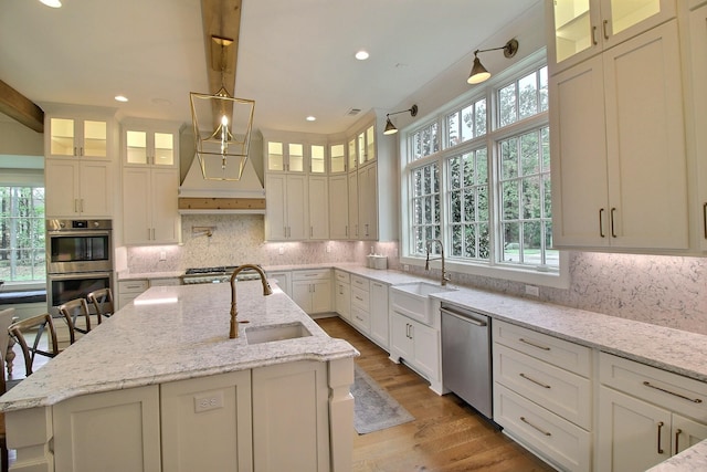 kitchen with stainless steel appliances, a center island with sink, sink, light stone countertops, and pendant lighting