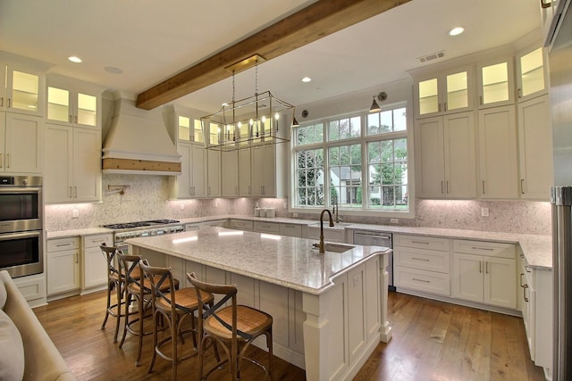 kitchen featuring appliances with stainless steel finishes, beam ceiling, hardwood / wood-style floors, a kitchen island with sink, and premium range hood