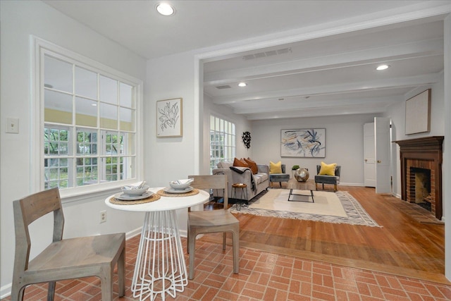 dining area featuring beamed ceiling, wood-type flooring, a healthy amount of sunlight, and a brick fireplace