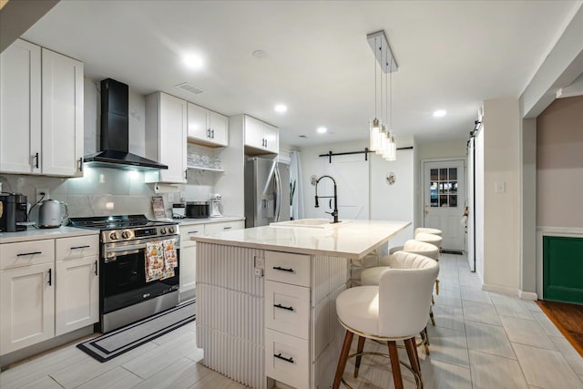 kitchen featuring wall chimney exhaust hood, a barn door, a kitchen island with sink, white cabinetry, and appliances with stainless steel finishes