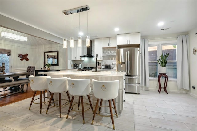 kitchen with stainless steel fridge, wall chimney range hood, hanging light fixtures, a breakfast bar, and white cabinets