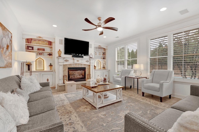 living room with built in features, ceiling fan, crown molding, and a tiled fireplace