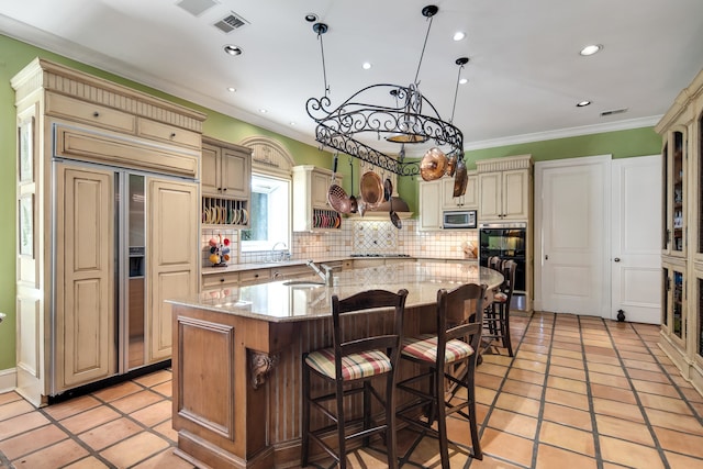 kitchen featuring light stone countertops, built in appliances, crown molding, cream cabinets, and an island with sink