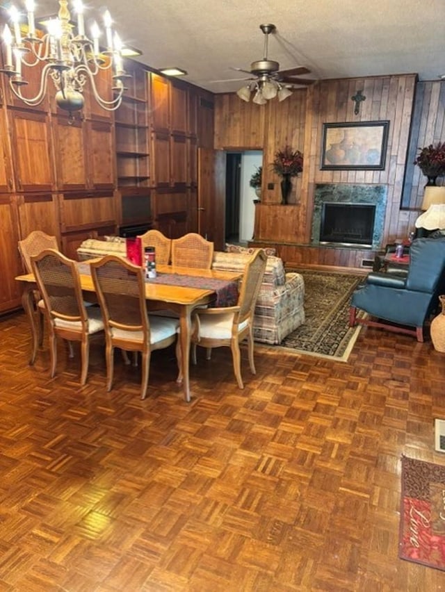 dining space with wood walls, ceiling fan with notable chandelier, dark parquet floors, a premium fireplace, and a textured ceiling