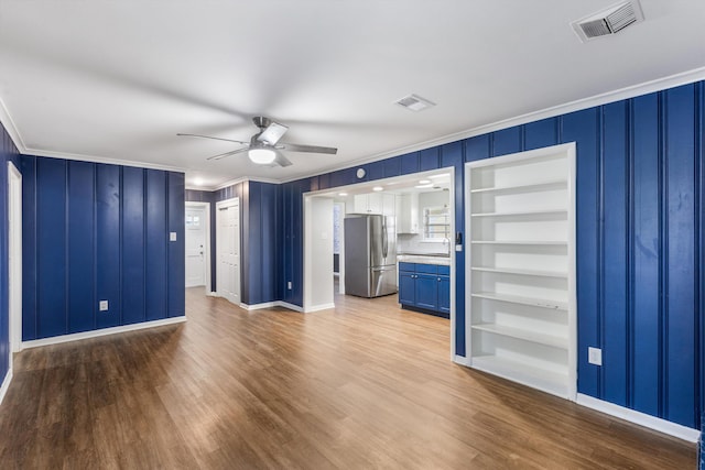 unfurnished living room featuring built in shelves, ceiling fan, wood-type flooring, and ornamental molding